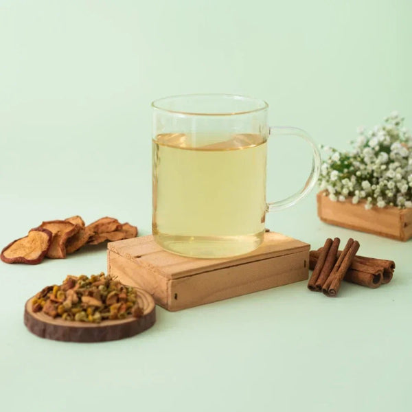 Caneca de vidro cheia de chá de camomila da marca Tea Shop, com ingredientes como maçã, canela e camomila fresca, dispostos em volta.